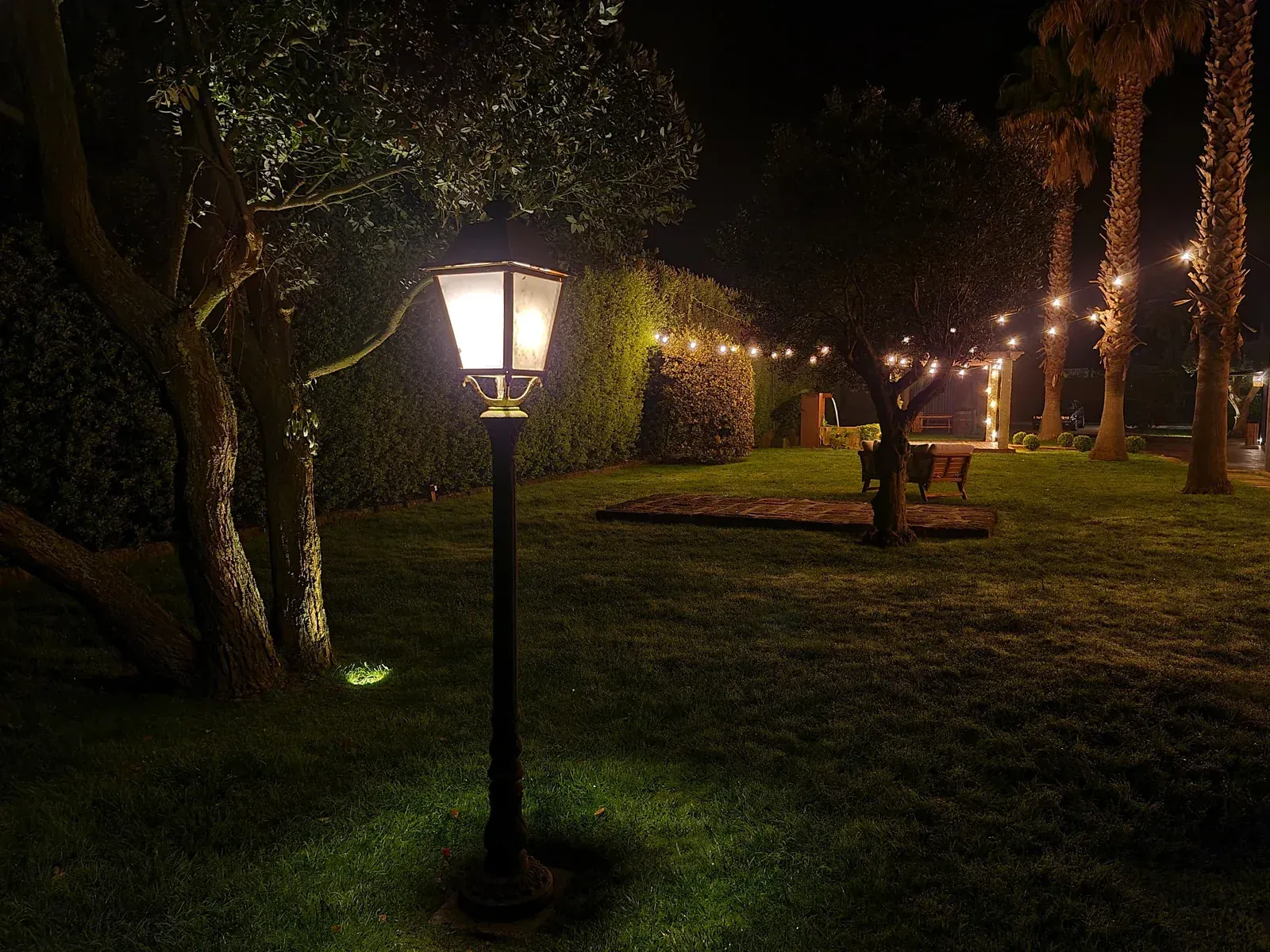 A lit vintage-style lamp post in a grassy garden at night, with string lights illuminating trees and a wooden bench.
