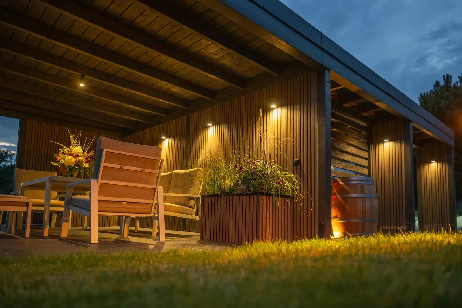 An outdoor patio with wood paneling, warm wall lighting, cozy lounge furniture, a large plant, and a wooden barrel.