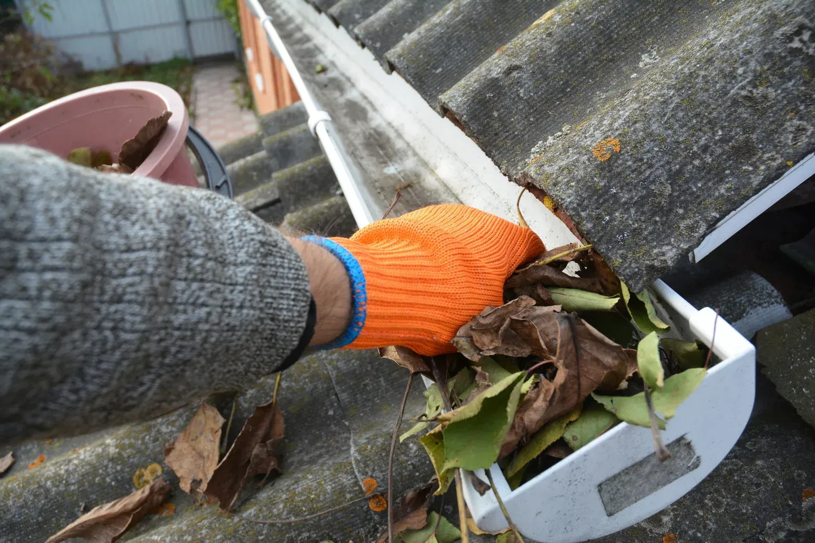 A person wearing a bright orange glove cleans dead leaves out of a white roof gutter.