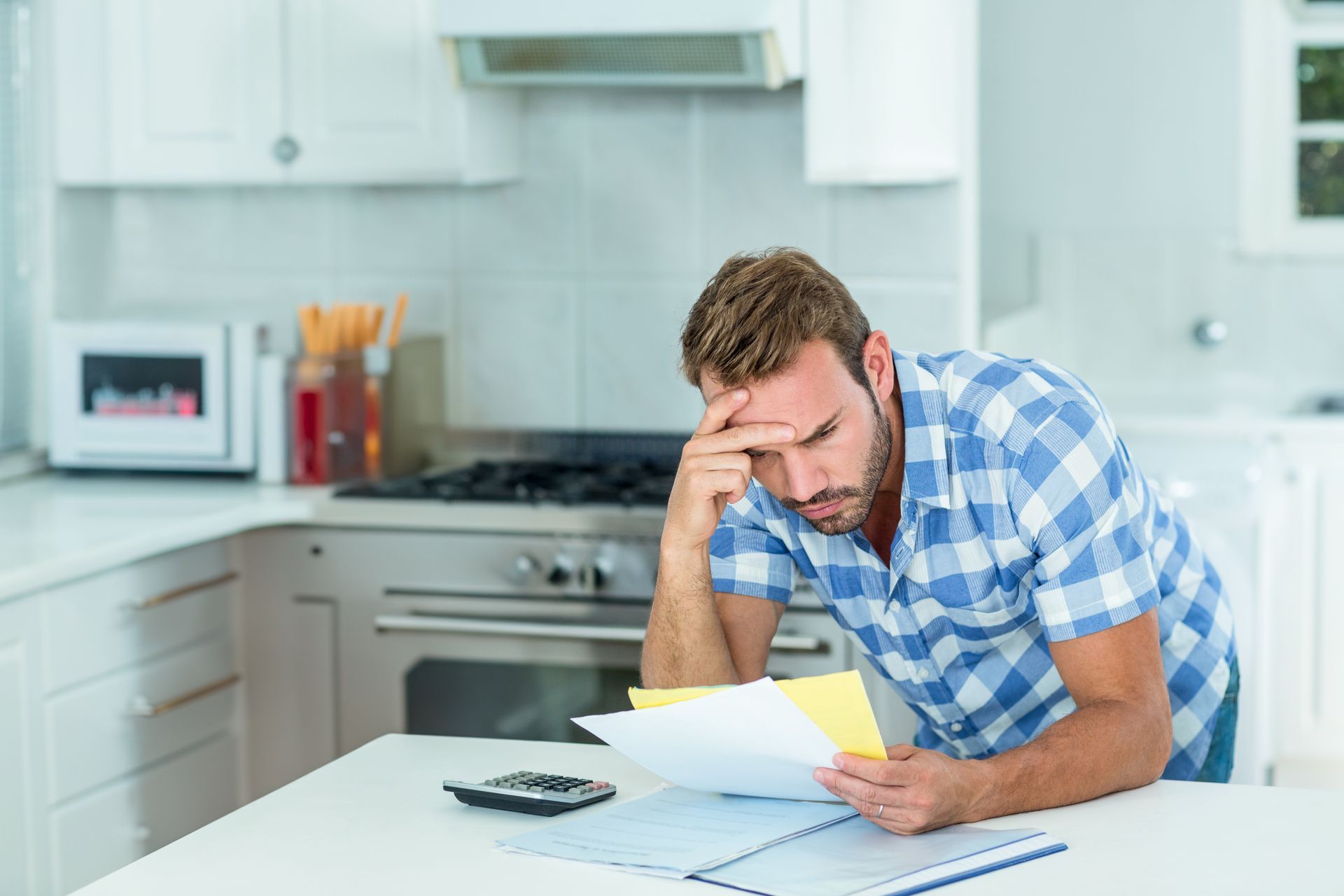 Man in kitchen, stressed, holding bills, calculator nearby.