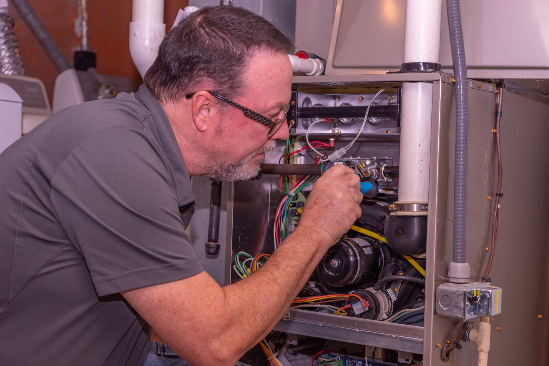 A man in glasses inspects a furnace, using a tool, in a utility room.