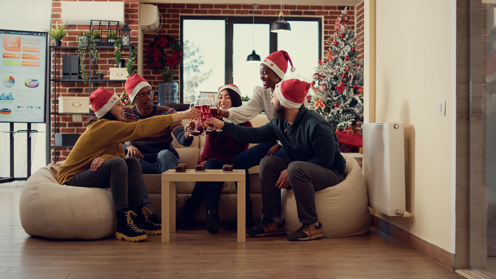 Group of friends in Santa hats toasting drinks, celebrating Christmas in a decorated office.