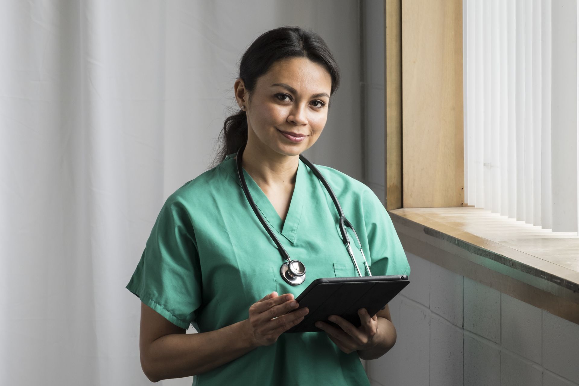 A nurse is standing in front of a window holding a tablet.
