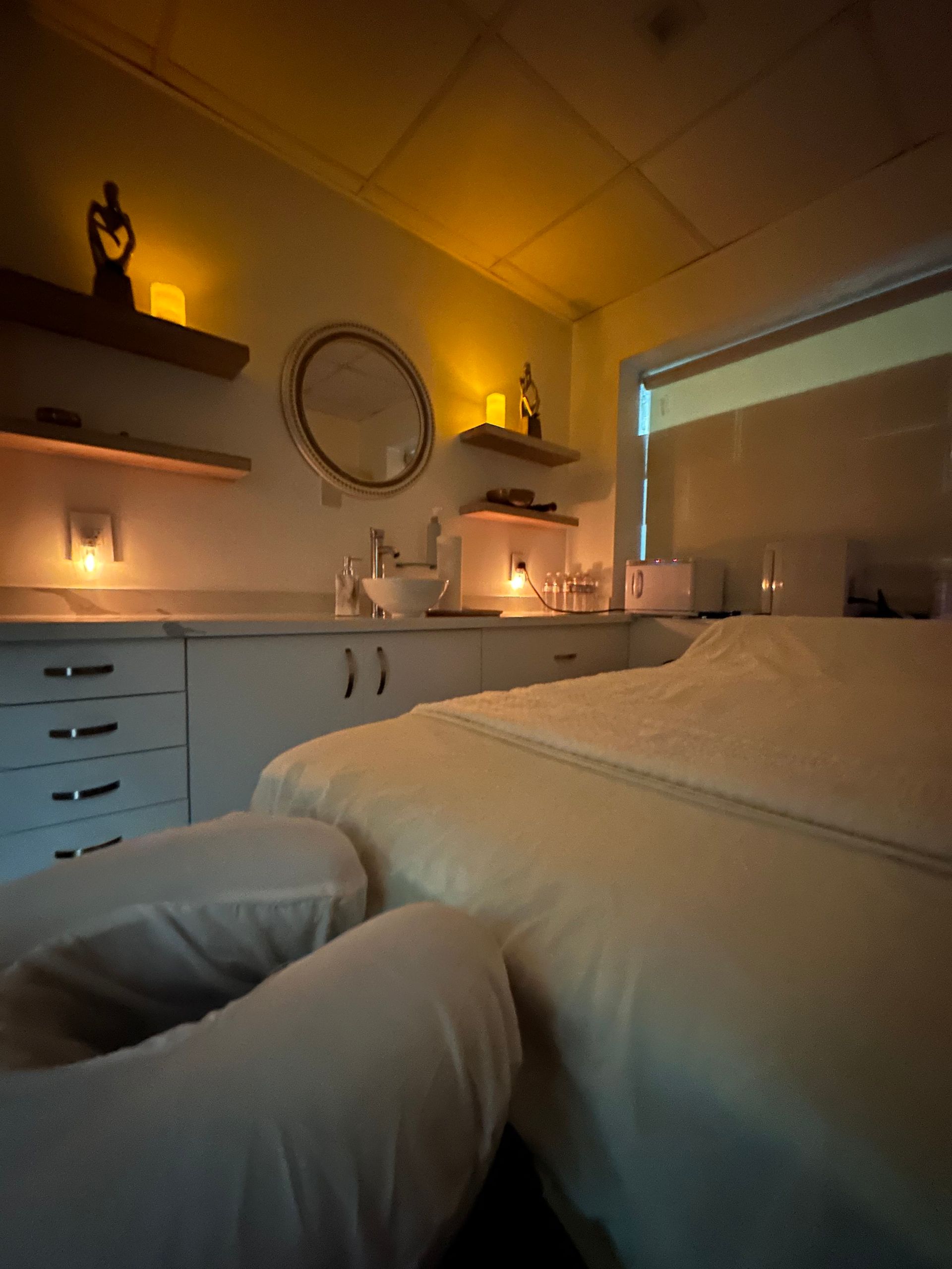 Cozy massage room with soft lighting, massage table, sink, and shelves with candles and decor.