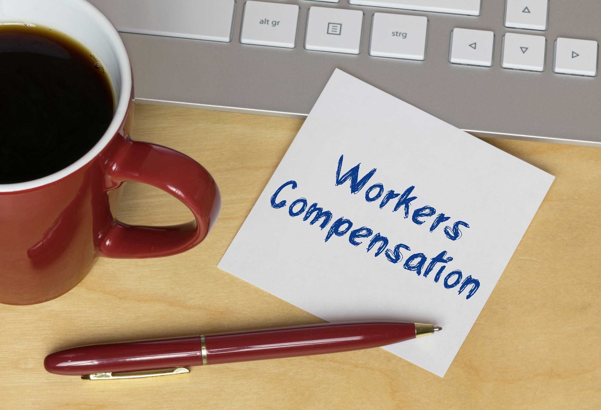 A note reading “Workers Compensation” sits beside a keyboard, coffee mug, and pen.