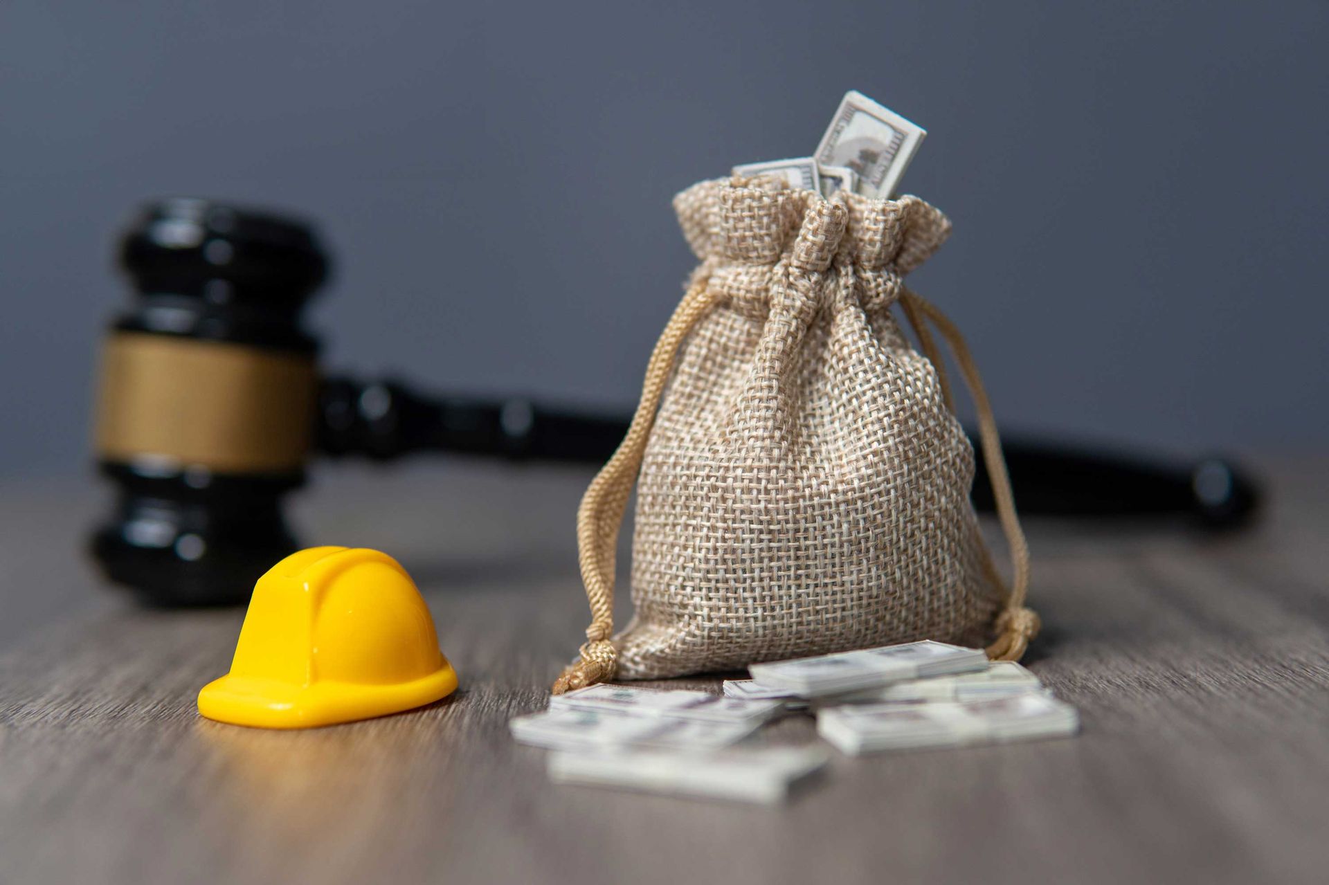 A small hard hat and a money bag sit on a table with a gavel blurred in the background.