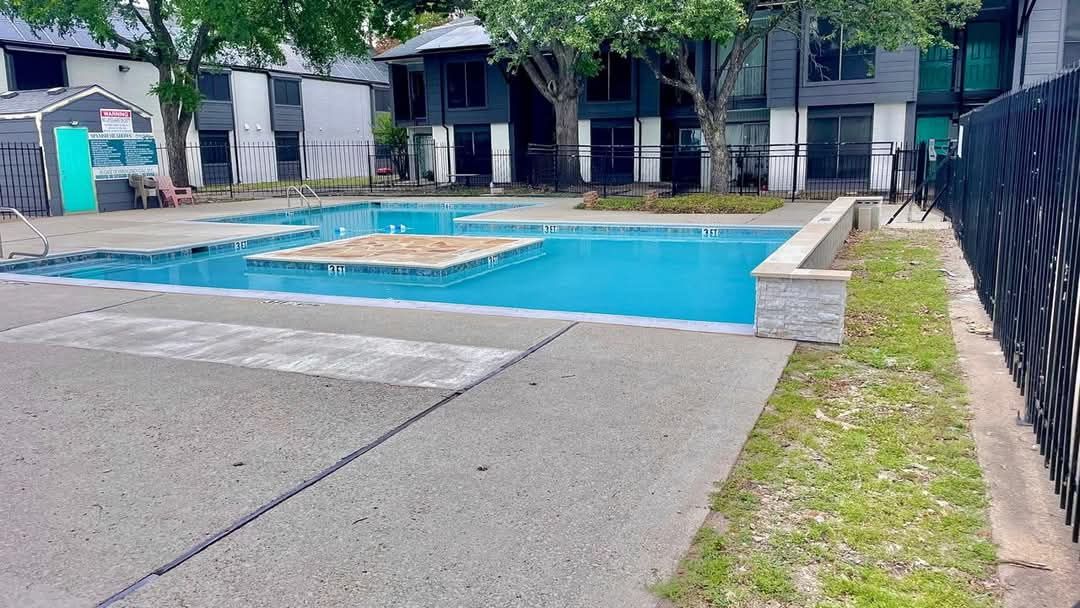 Pool surrounded by concrete with buildings in the background; grassy area along a black fence.