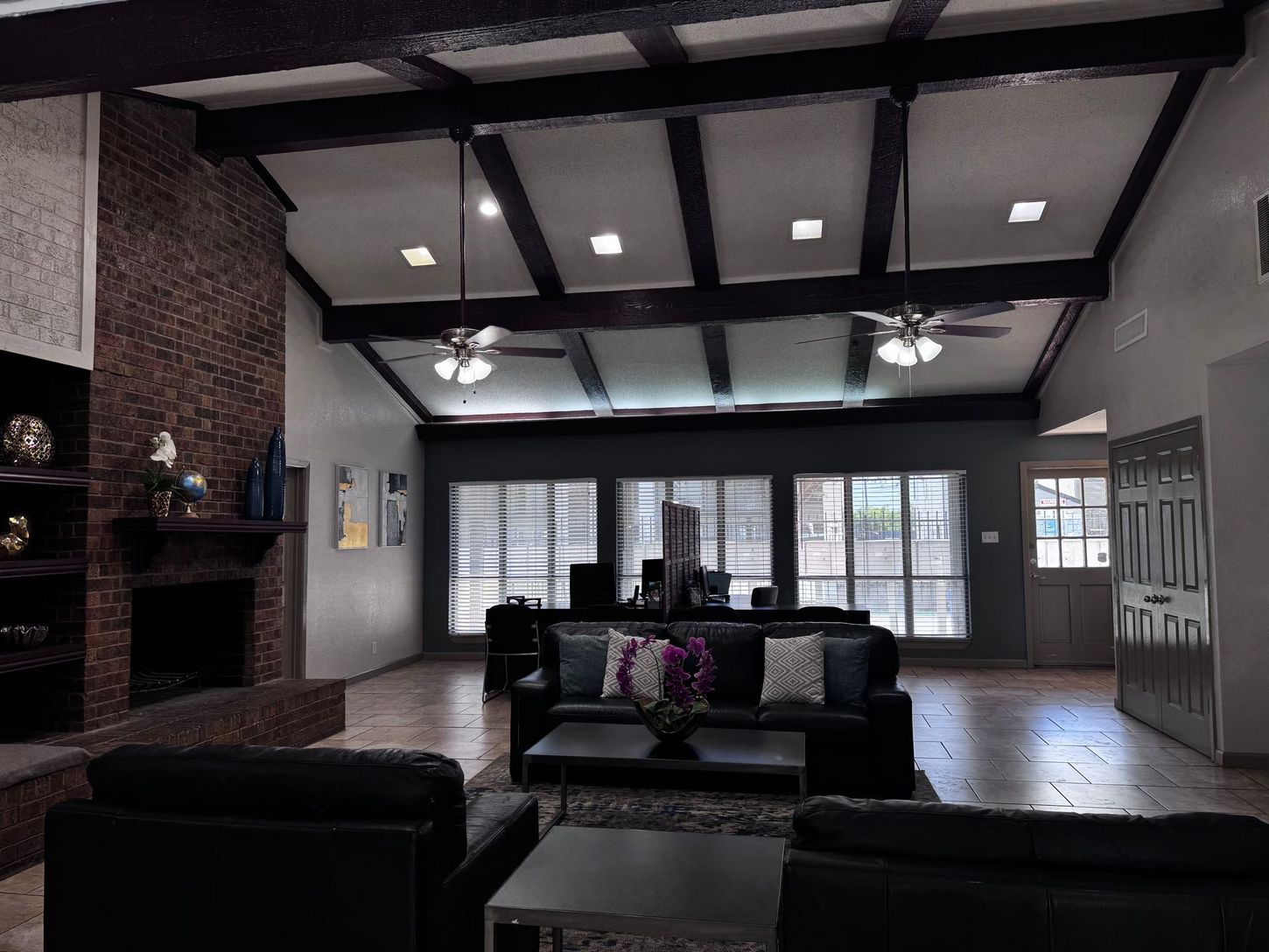 Living room with high ceiling, exposed beams, fireplace, black furniture, and large windows.
