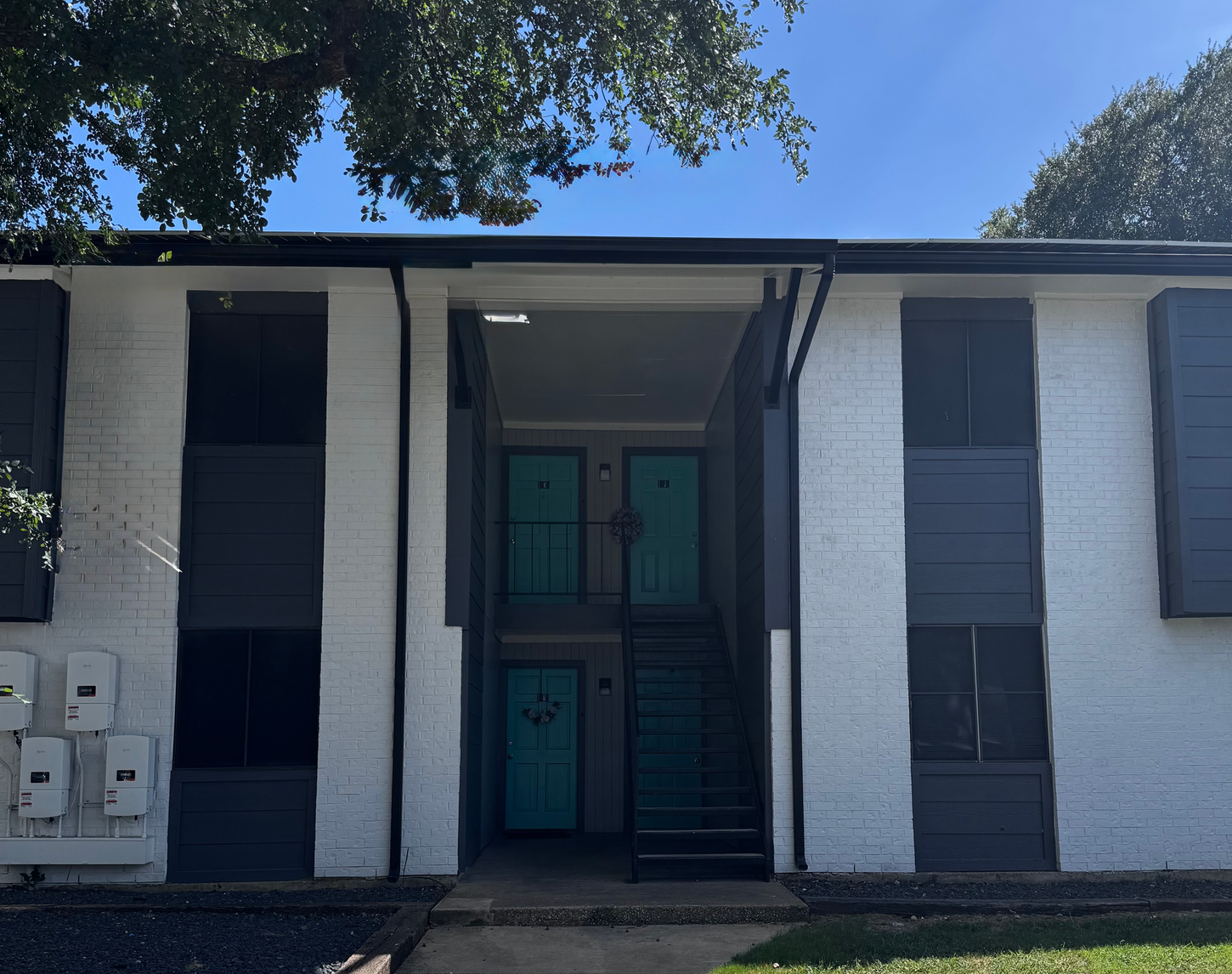 Two-story apartment building with white brick, blue-gray accents, teal doors, and black shutters.