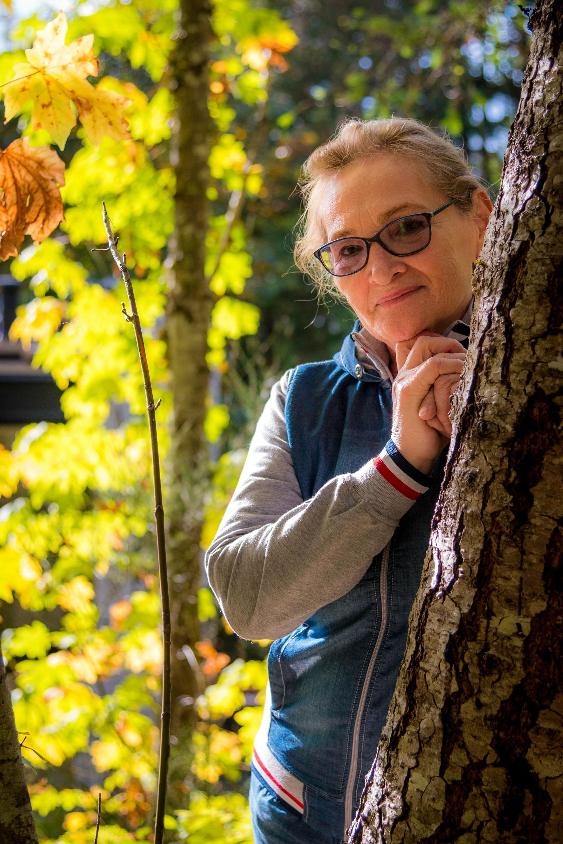 Woman with glasses peeks from behind a tree, fall foliage in the background.
