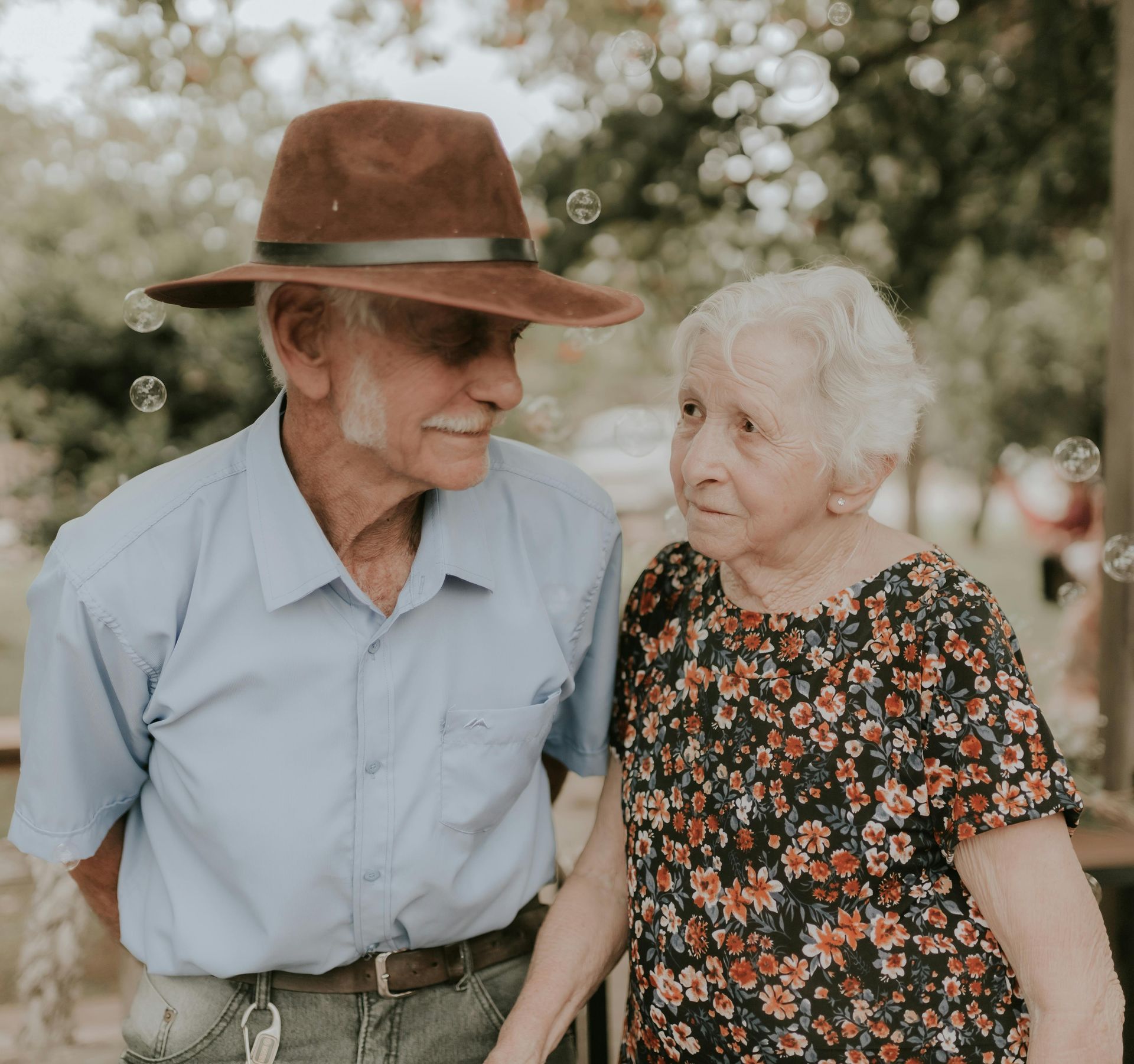 Elderly couple smiles lovingly at each other; man in brown hat and blue shirt, woman in floral top. Outdoor setting.
