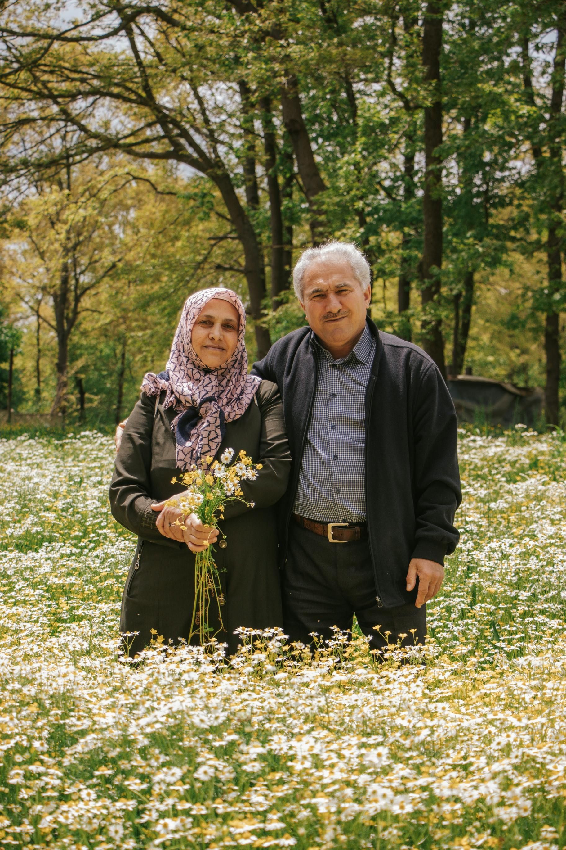 Elderly couple standing in a field of white flowers, smiling. Woman wears headscarf, holding flowers. Trees in background.