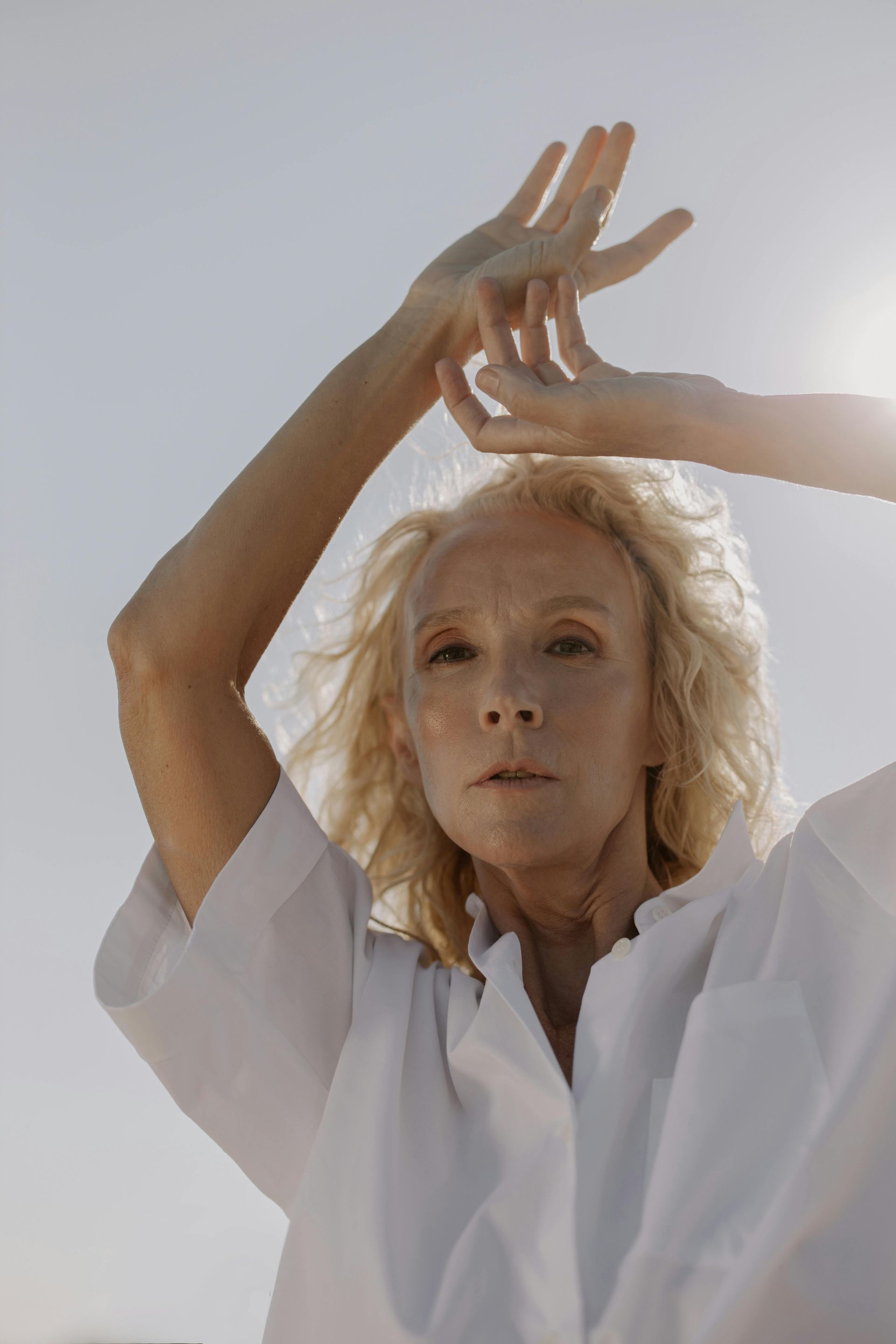 Woman with blonde hair in white shirt, arms raised. Outdoors in sunlight.