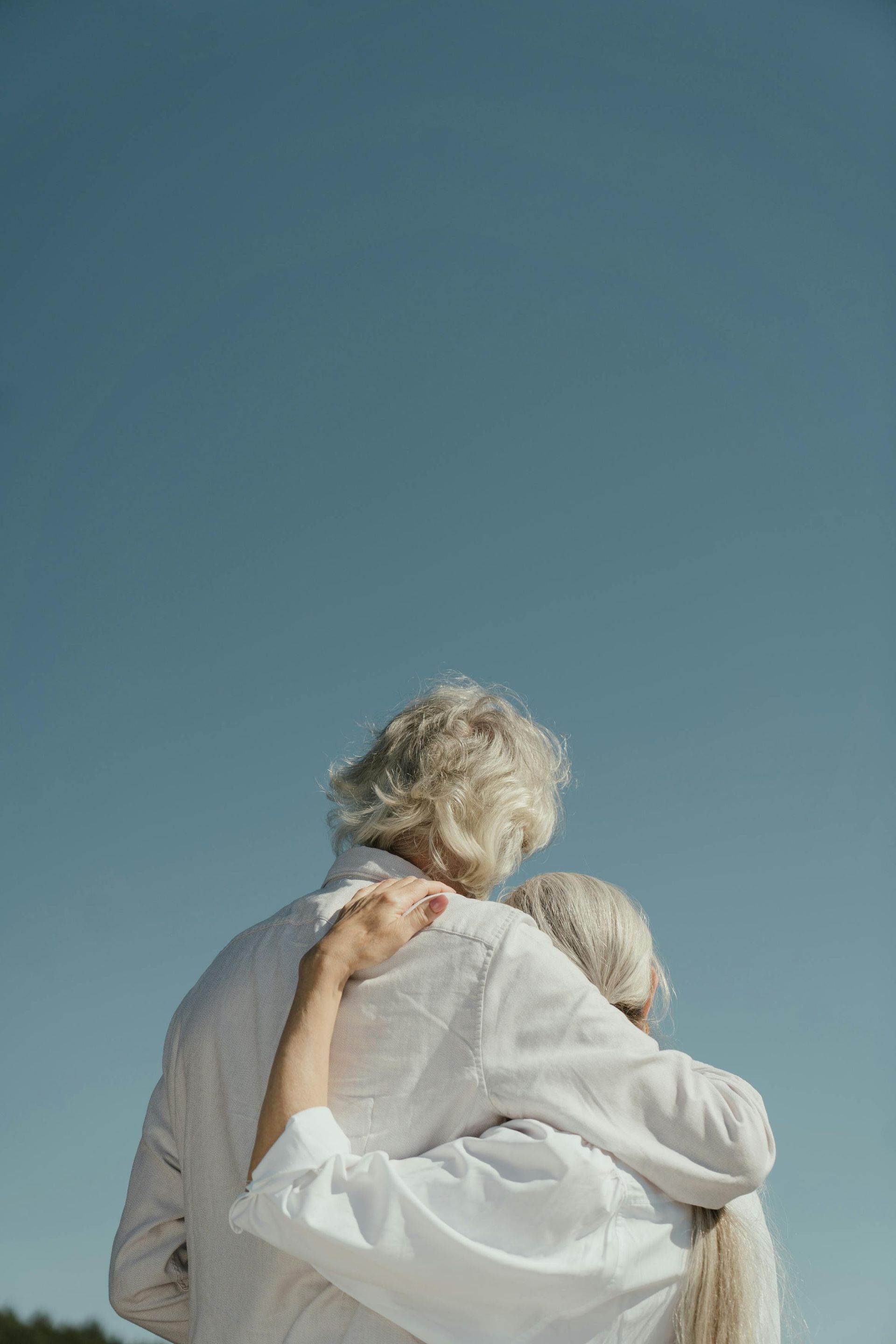 Elderly couple embracing, backs to the camera, against a clear blue sky.