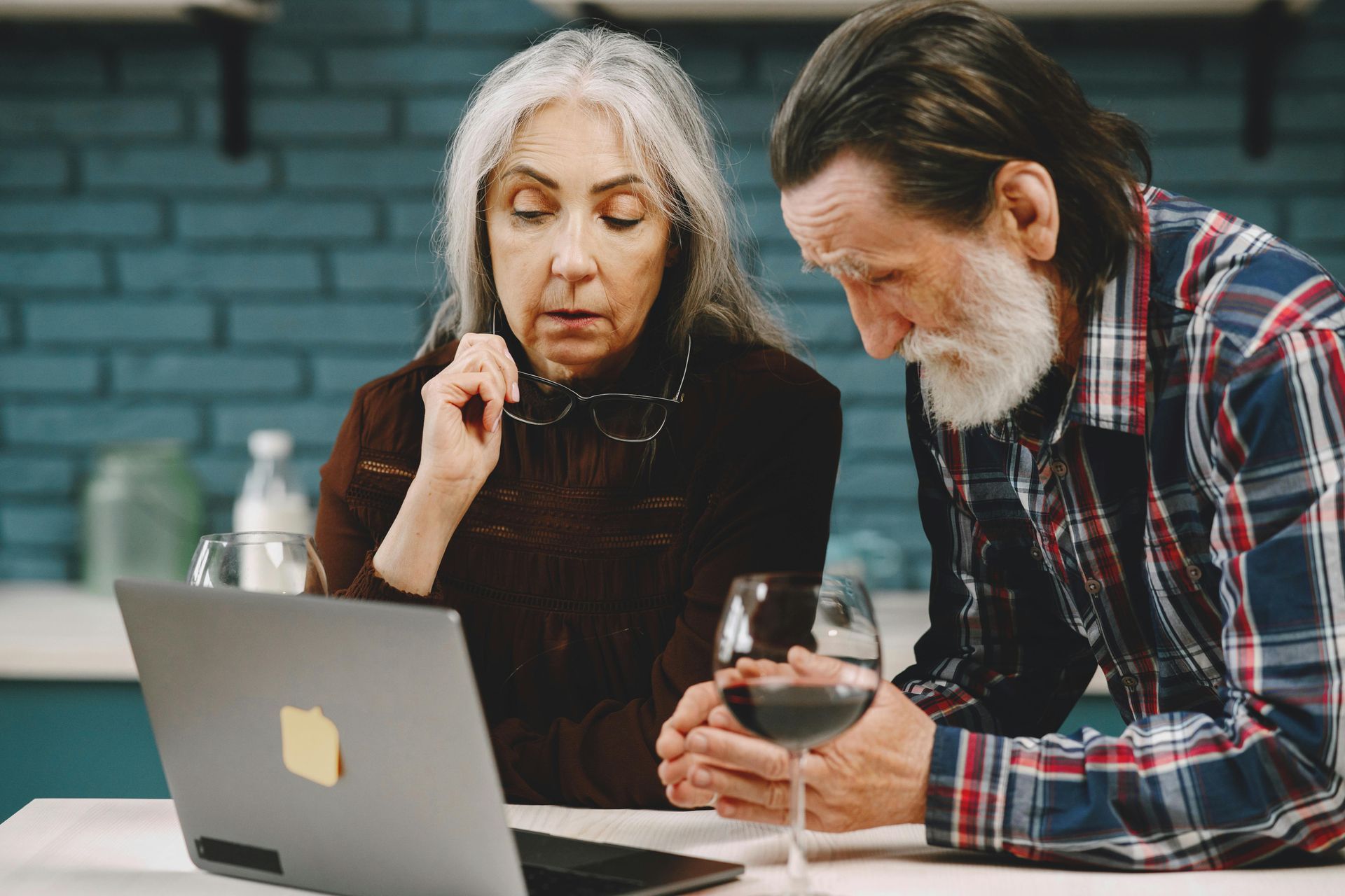 Older couple looking at a laptop together. The woman gestures thoughtfully; the man holds a wine glass.