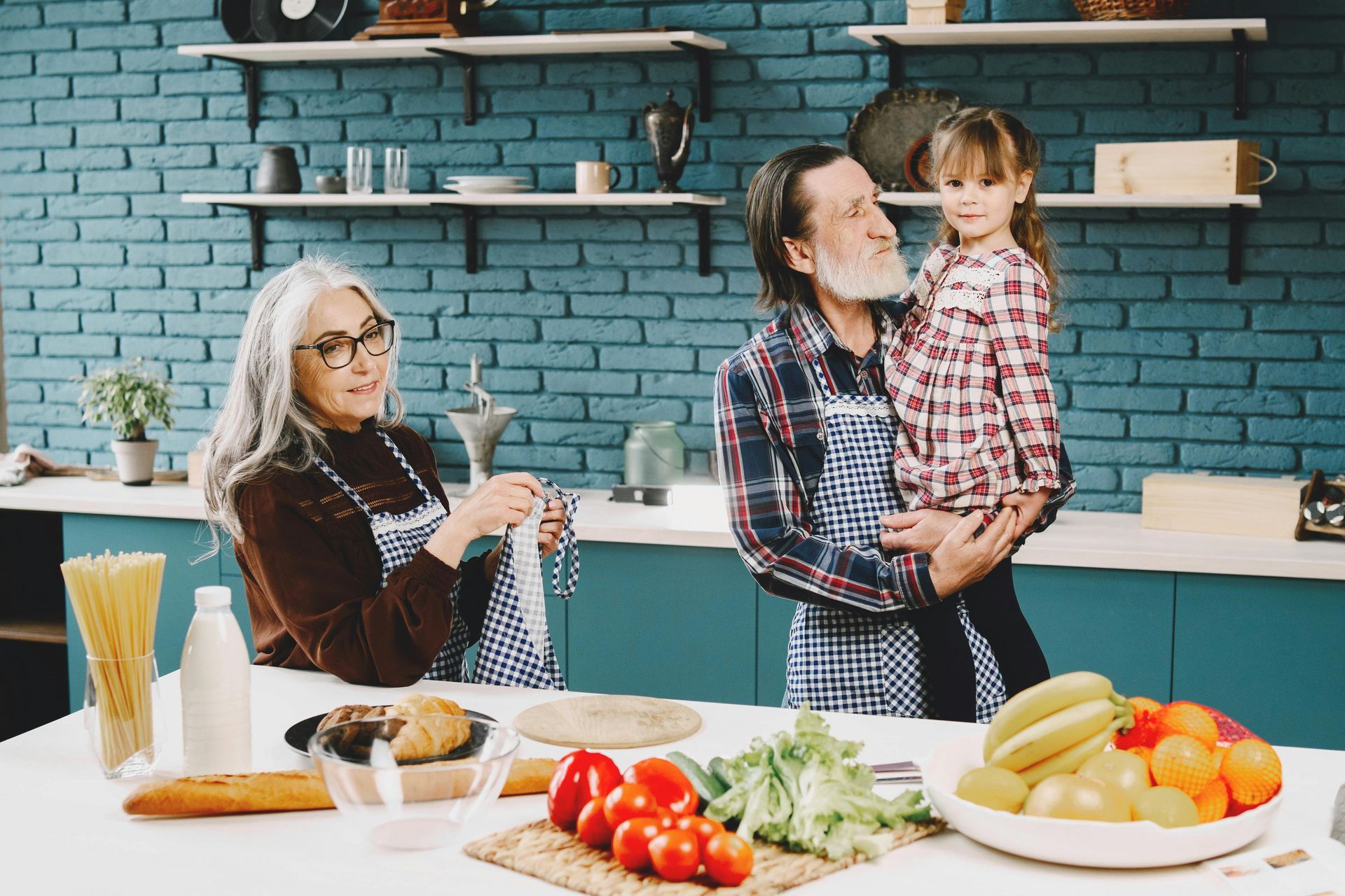 Grandparents and granddaughter in a kitchen. Woman knitting, man holding girl. Food prep on counter.