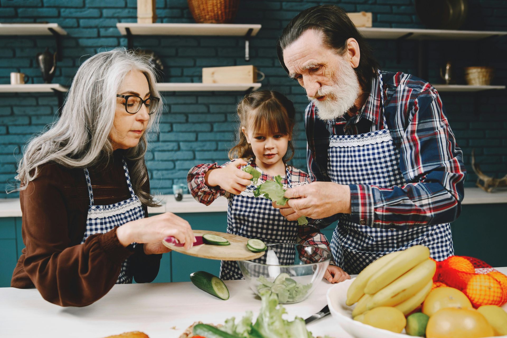 Grandparents and child prepare food in kitchen. They cut vegetables while wearing aprons, with fruit nearby.