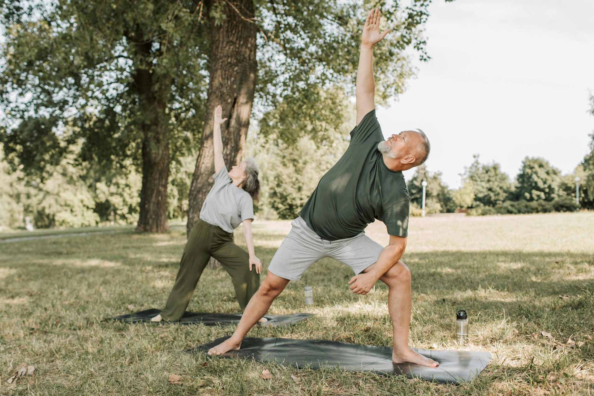 Two people doing yoga outdoors: one in a triangle pose, the other in a reverse warrior pose, in a sunny park.