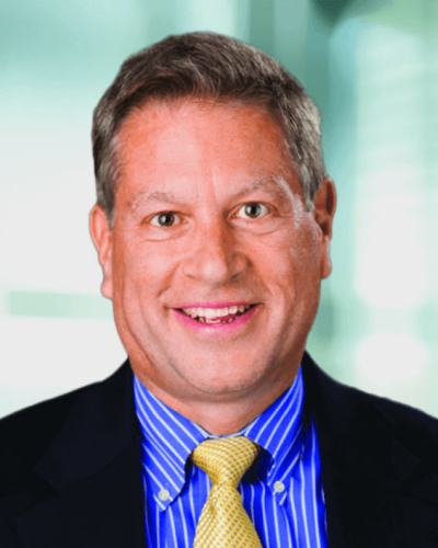 Man smiling in a blue striped shirt and yellow tie, wearing a dark suit jacket, business setting.