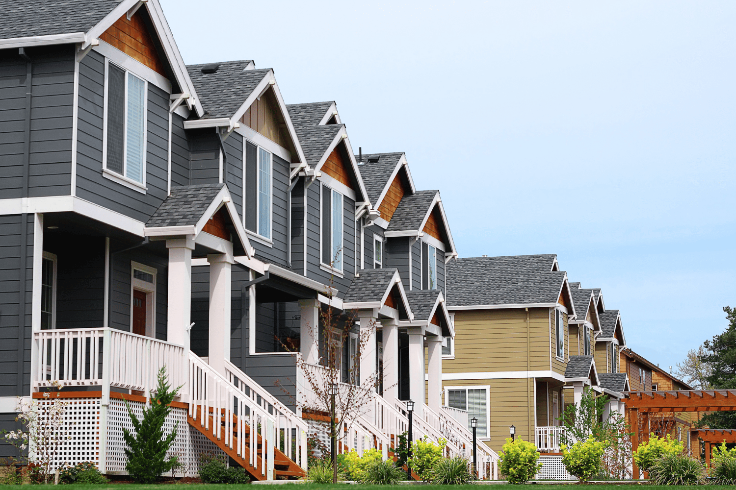 Row of townhouses with gray and tan siding, white trim, and small front porches, under a light blue sky.