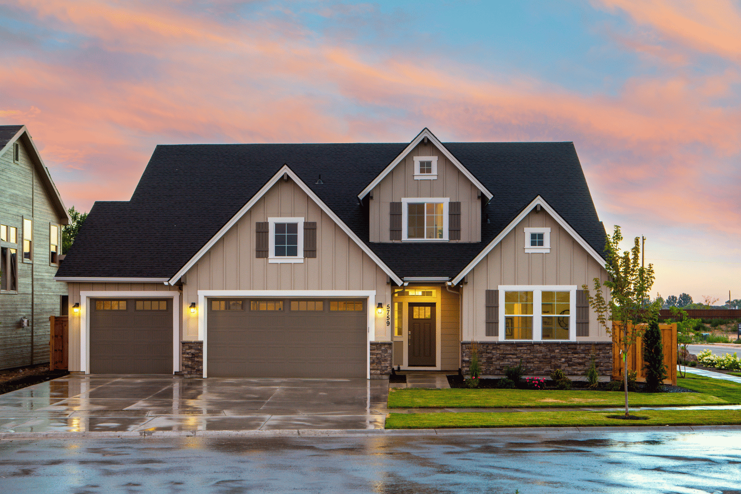 Beige two-story house with black roof and stone accents, two-car garage, on a lawn with a cloudy sunset.