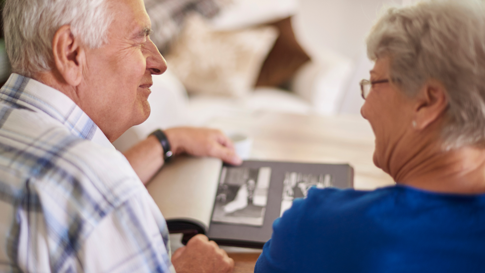 Older couple looking at a photo album, smiling. Indoors, natural light, soft focus.