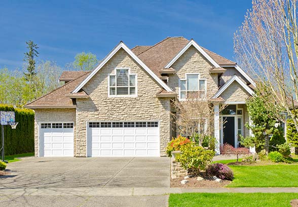 Two-story beige stone house with brown roof, white garage doors, green lawn, and blue sky.