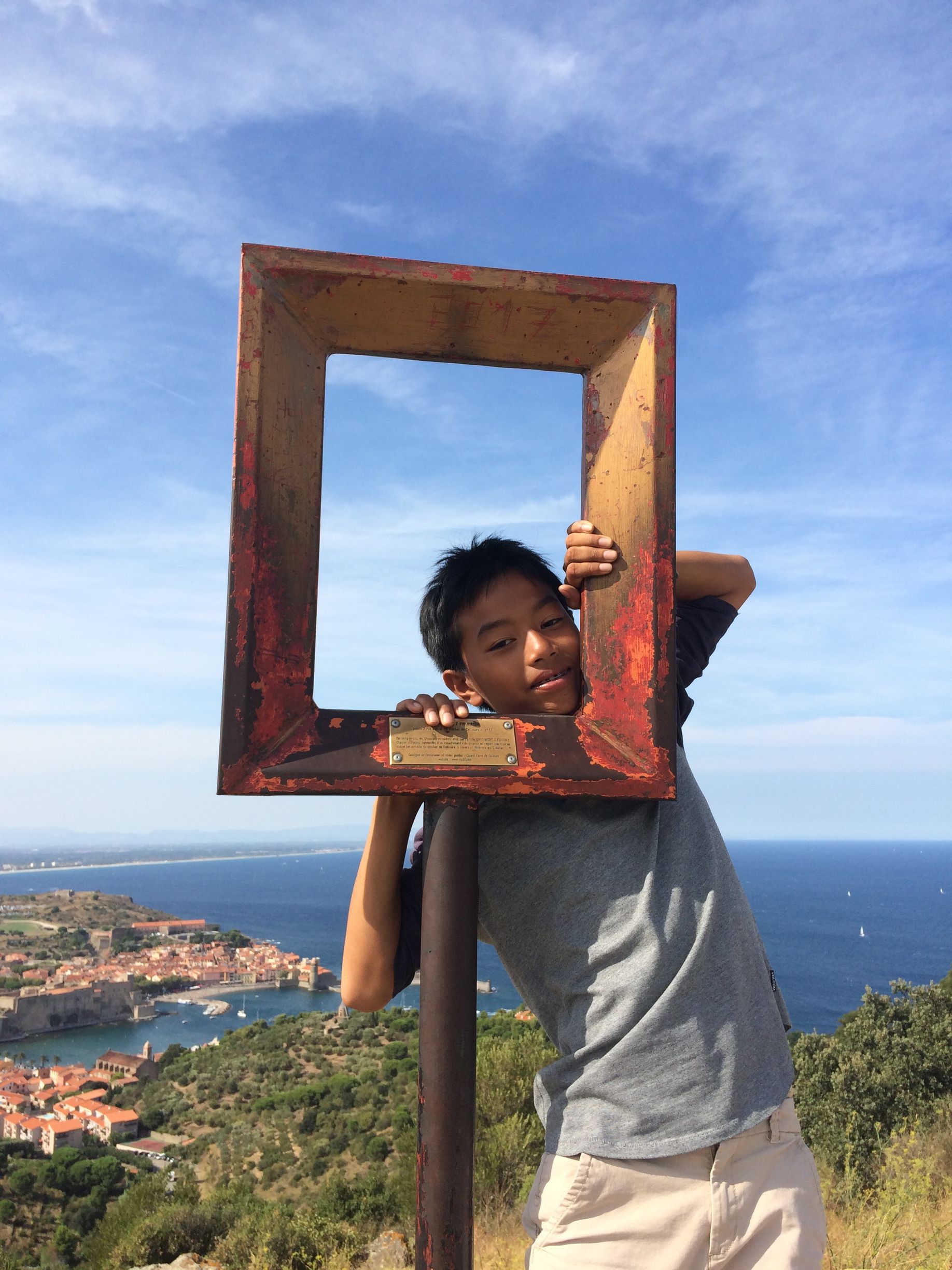Une personne pose à l'intérieur d'un cadre rouillé, sur une colline surplombant une ville. Ciel bleu et mer en arrière-plan.