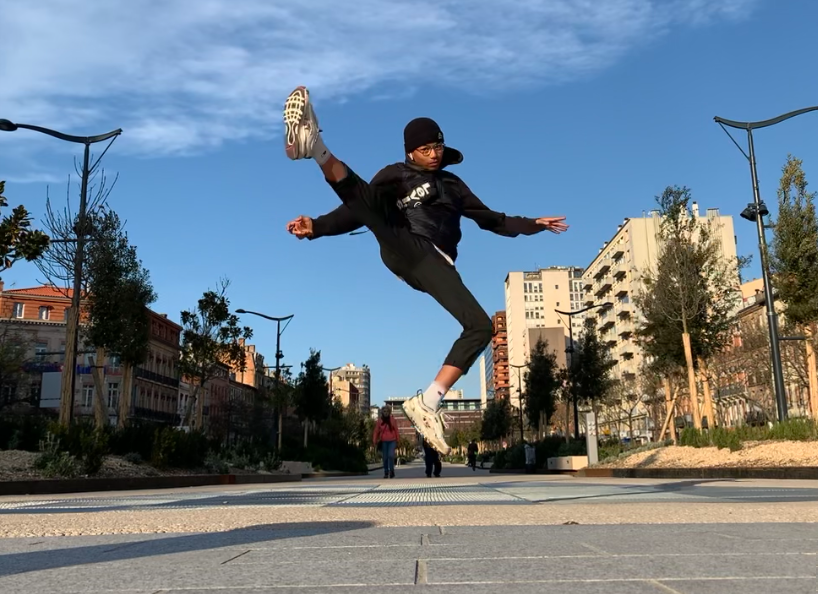 Une personne saute dans les airs, exécutant un mouvement de danse dans une rue. Bâtiments, arbres et ciel bleu.