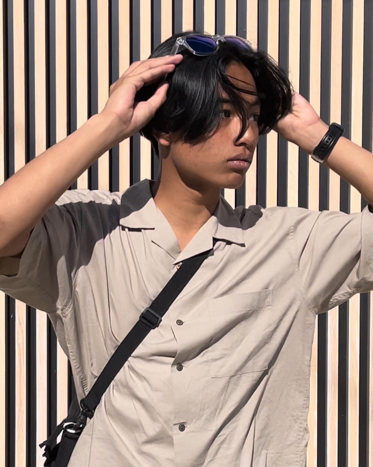 Un homme en chemise beige et lunettes de soleil, se recoiffant, avec un sac bandoulière noir, devant un mur rayé.