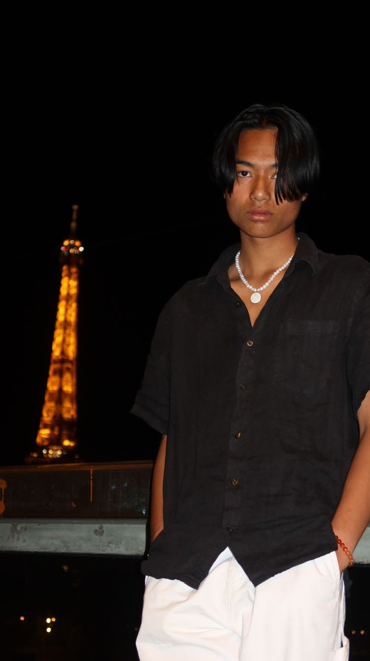 Jeune personne en chemise noire et pantalon blanc, la Tour Eiffel illuminée en arrière-plan.