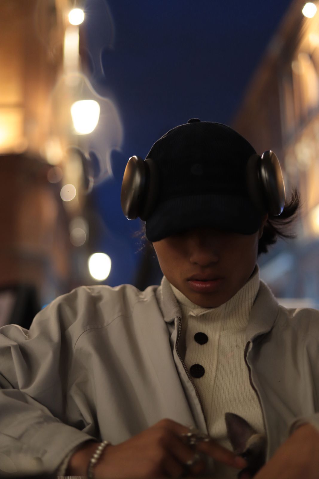 Un homme en chemise beige et lunettes de soleil, se recoiffant, avec un sac bandoulière noir, devant un mur rayé.