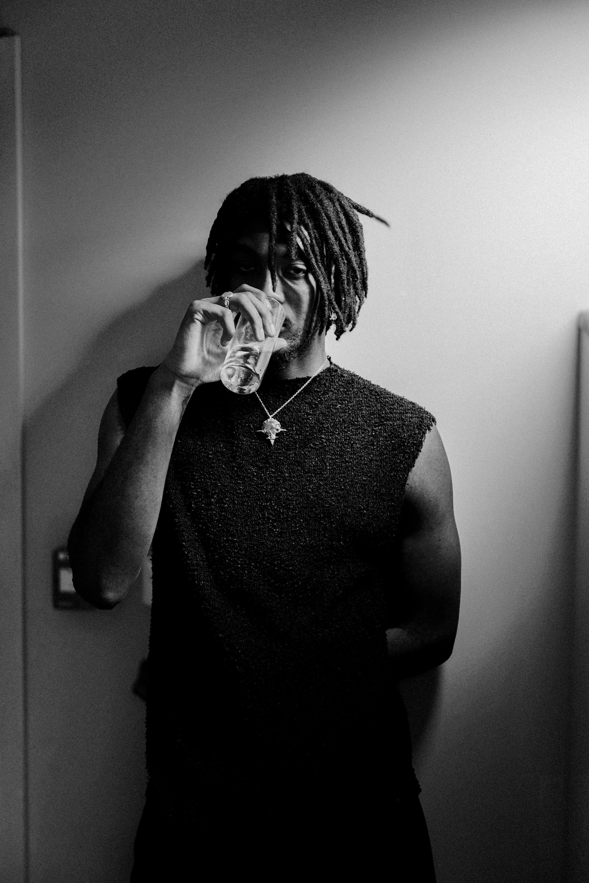 Personne buvant dans un verre, portant un débardeur et des dreadlocks, appuyée contre un mur.