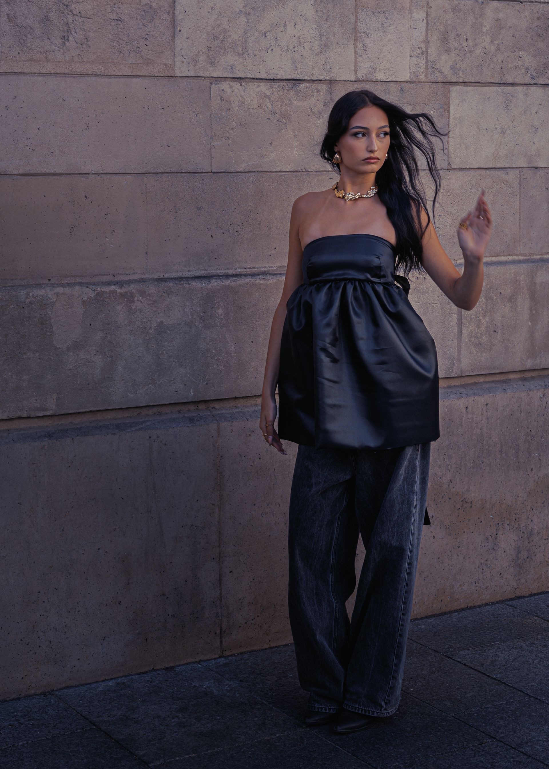 Une femme en haut noir et jean fait signe de la main devant un mur de pierre.