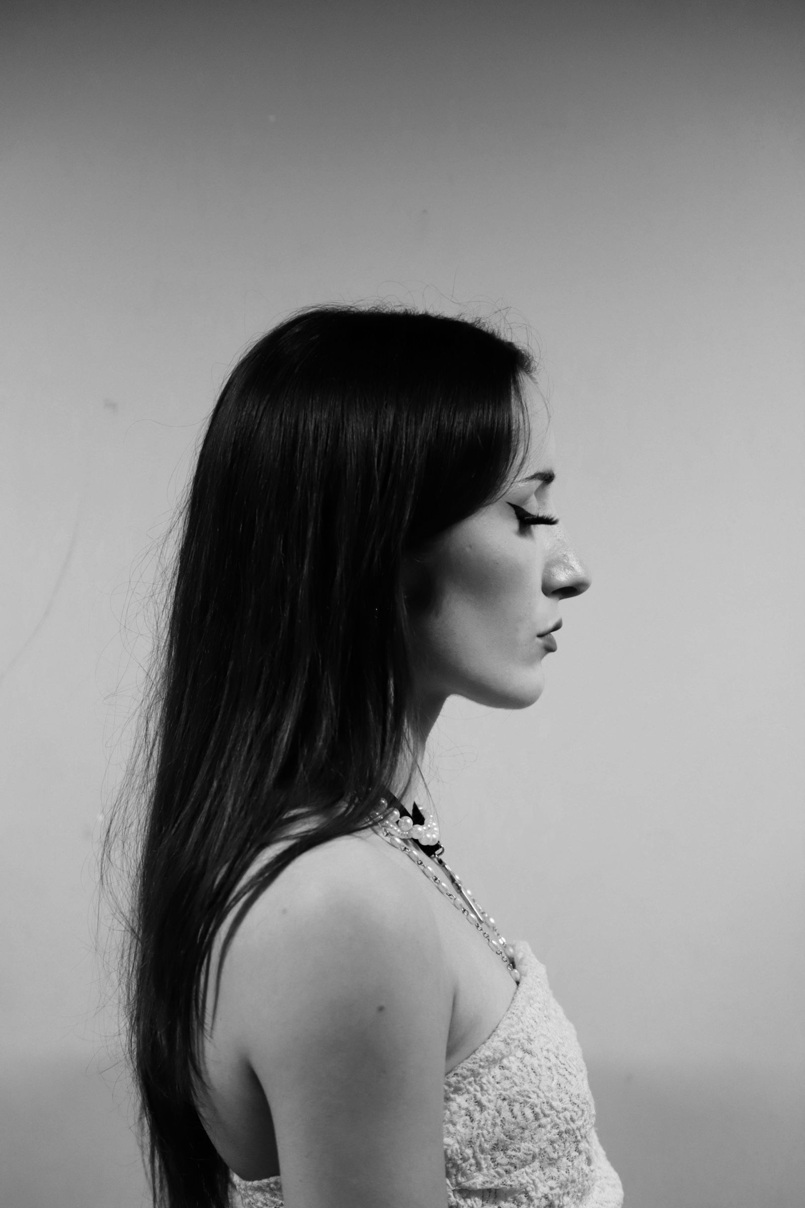 Profil d'une femme ; longs cheveux noirs, portant un haut en dentelle blanche, piercing à la lèvre, sur fond uni.