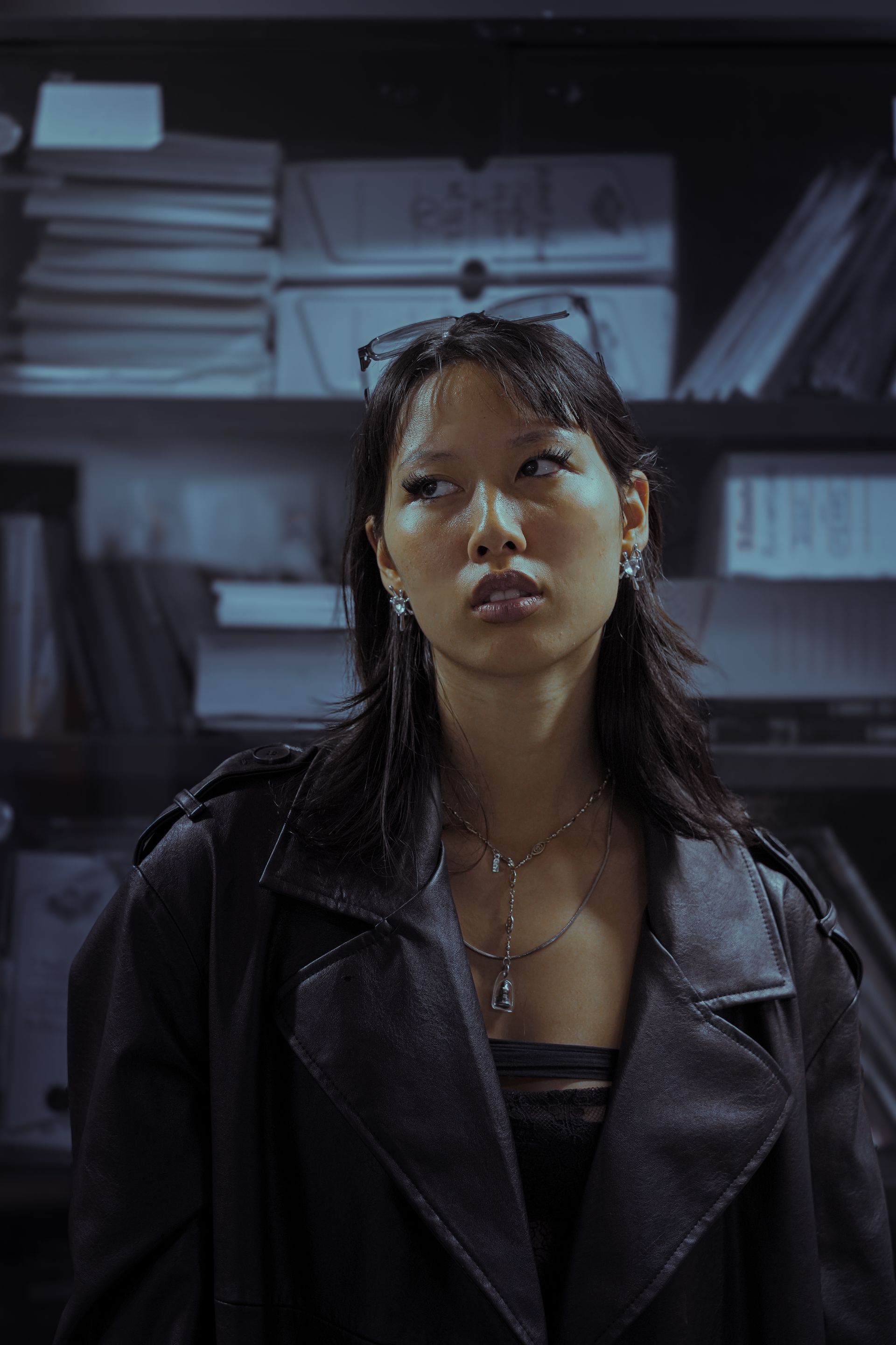Femme en manteau de cuir regardant vers le haut, devant une bibliothèque.