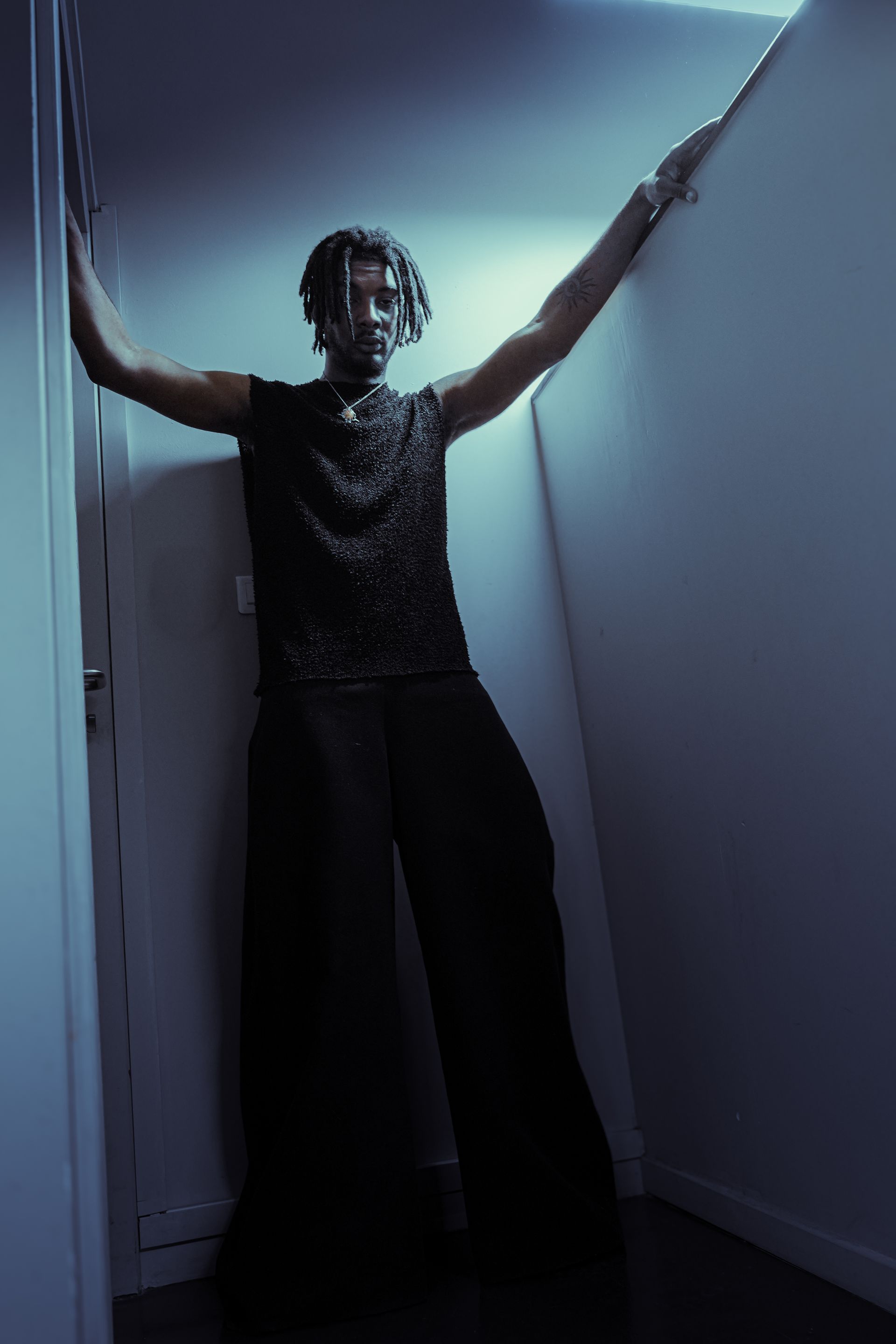 Une personne appuyée contre un mur dans un couloir faiblement éclairé, les bras écartés. 