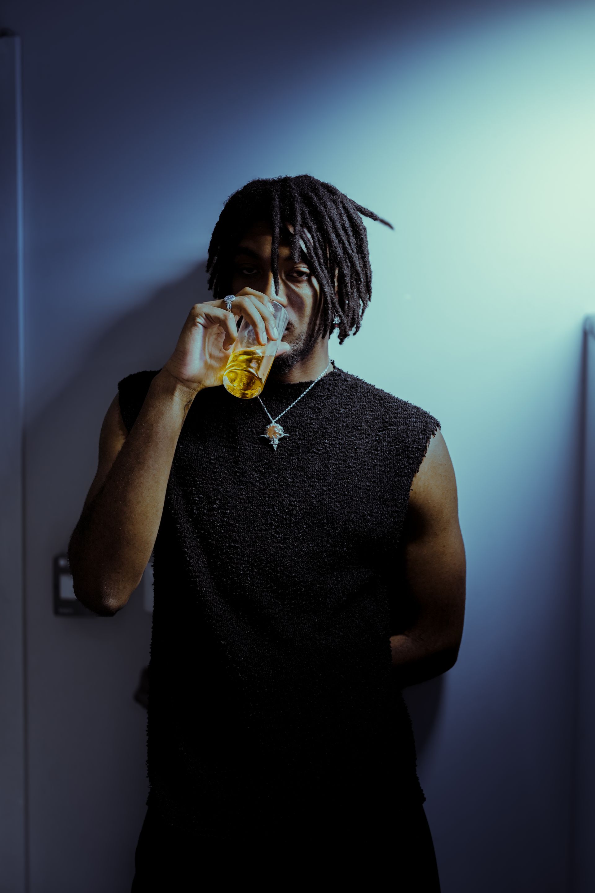 Une personne boit dans un verre, appuyée contre un mur. Elle porte un débardeur noir et a des dreadlocks.