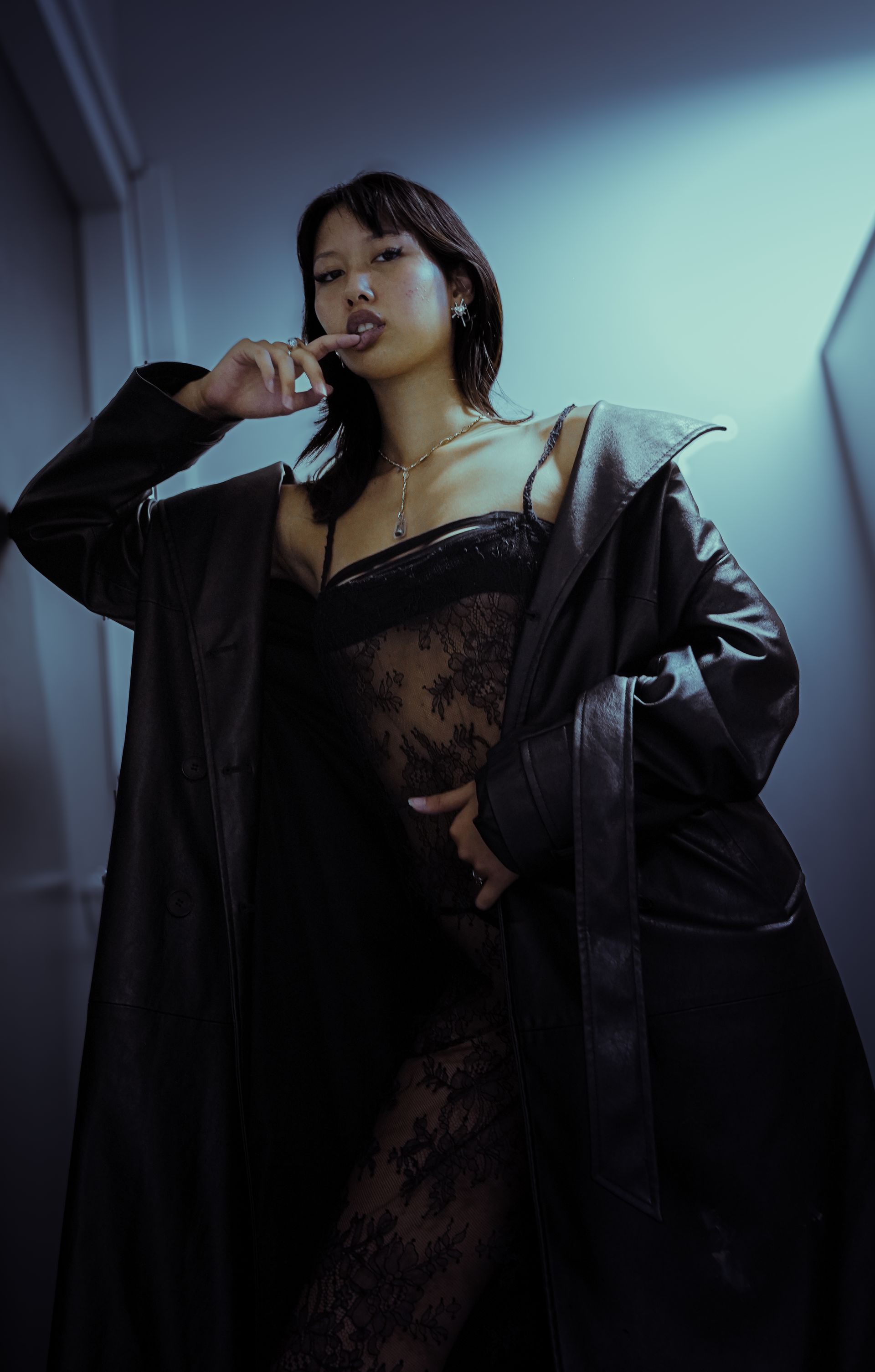 Femme en nuisette de dentelle noire et manteau de cuir, posant dans l'embrasure d'une porte, regardant l'objectif.