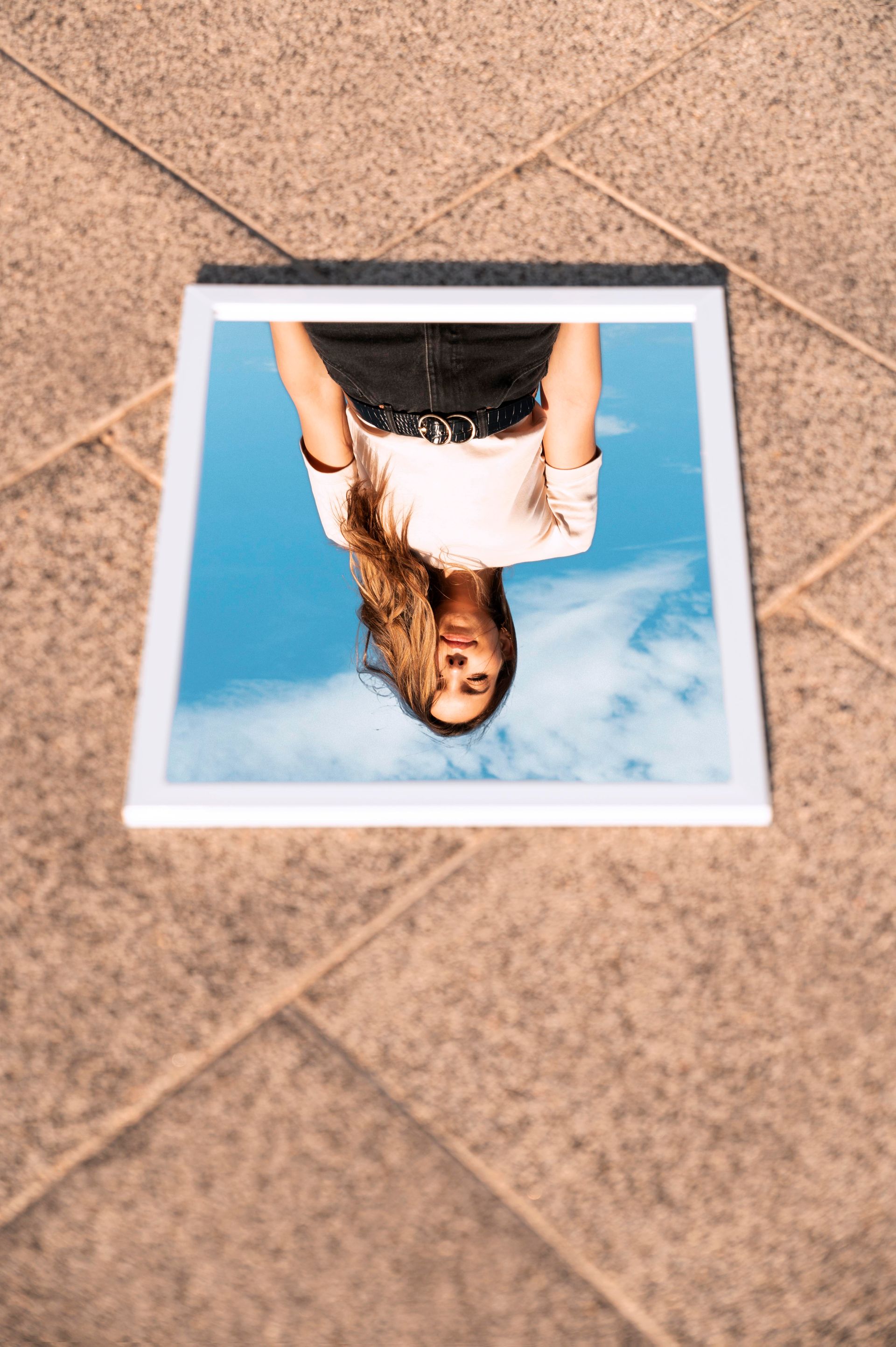 Une femme en haut blanc et jupe noire se reflète dans un miroir sur un ciel bleu, sur un sol en pierre.