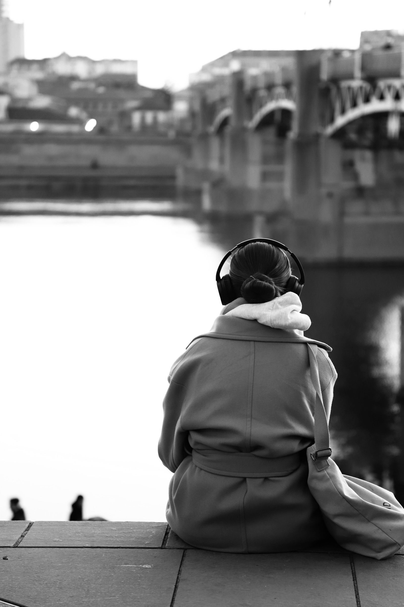 Une femme avec des écouteurs est assise près d'un pont, surplombant l'eau. Noir et blanc.