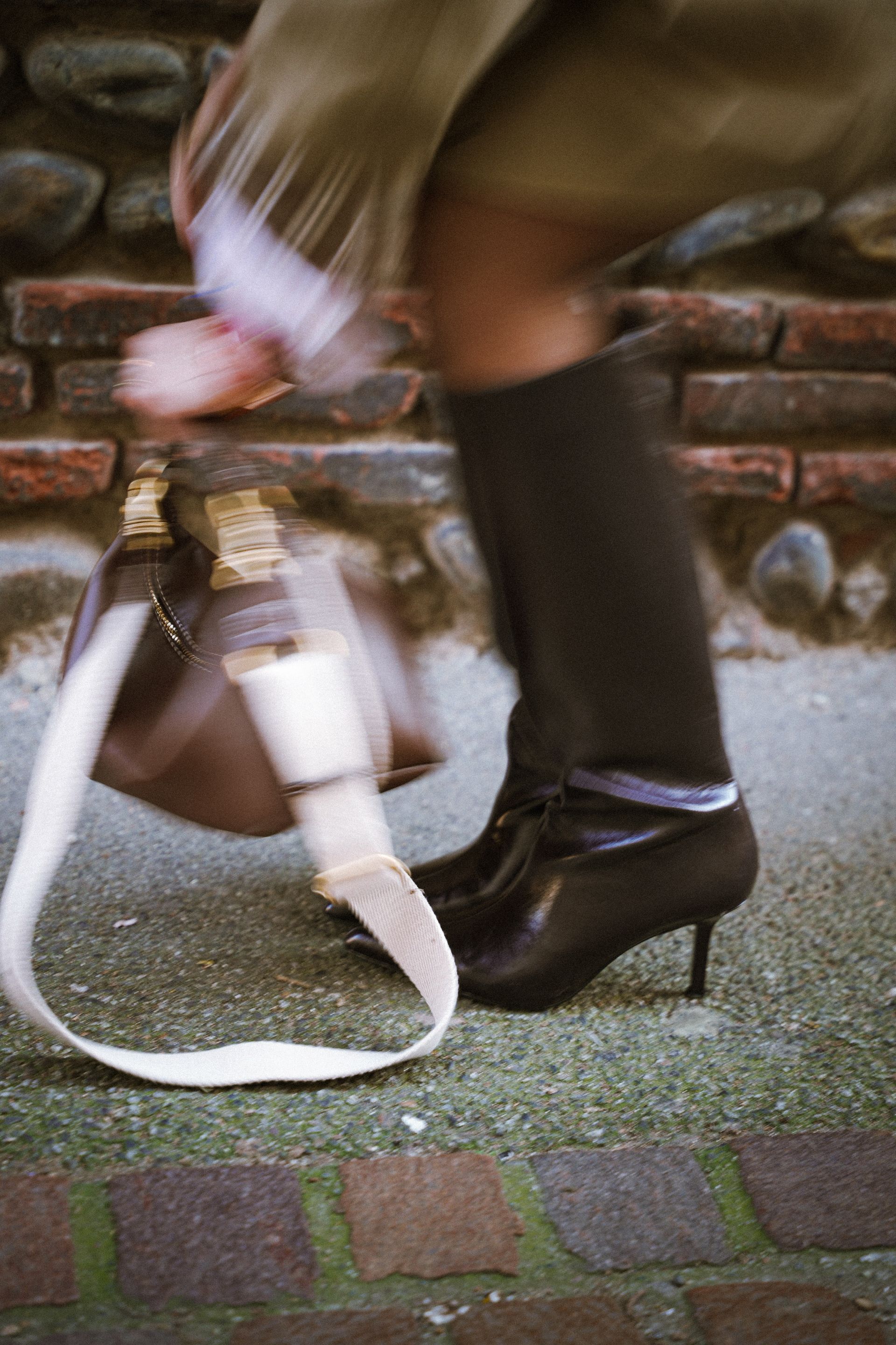 Personne portant des bottes noires et un manteau beige, attrapant un sac à main beige sur un chemin pavé.