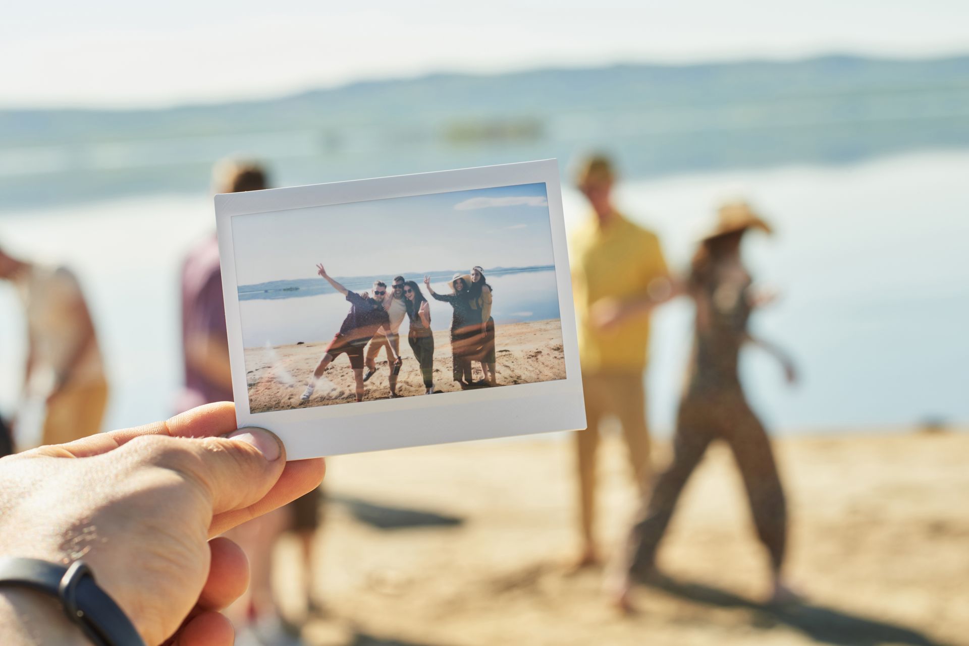 Une main tient une photo Polaroid de trois personnes sur une plage, avec d'autres personnes en arrière-plan. 