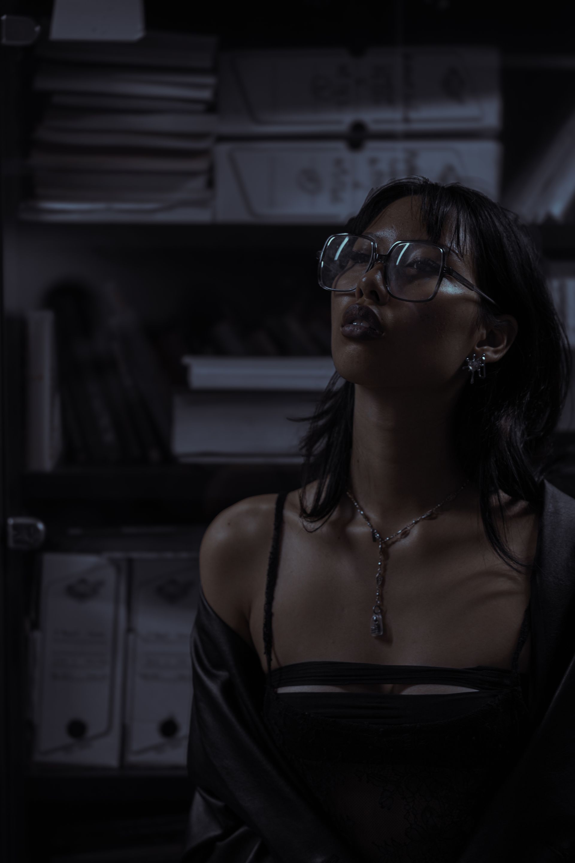 Femme en lunettes et débardeur près d'une étagère ; le regard levé, l'air pensif. Ambiance sombre et mystérieuse.