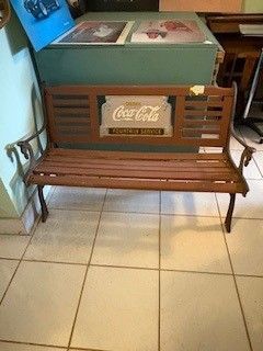 Wooden bench with Coca-Cola sign, in front of a green cooler, on tiled floor.