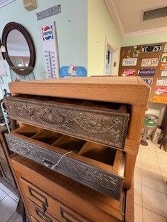 Wooden chest of drawers with two open drawers, showing metal decorative trim, in a cluttered room.