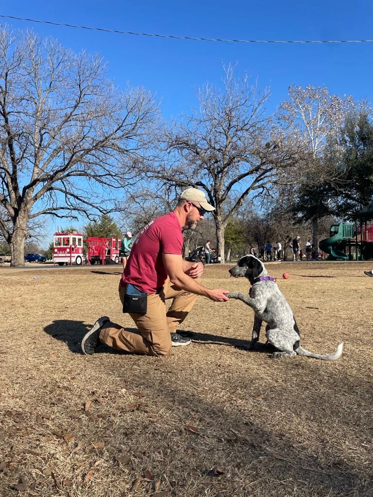 Man in brown pants kneels, giving a paw to a dog on a grassy field; fire truck visible in background.