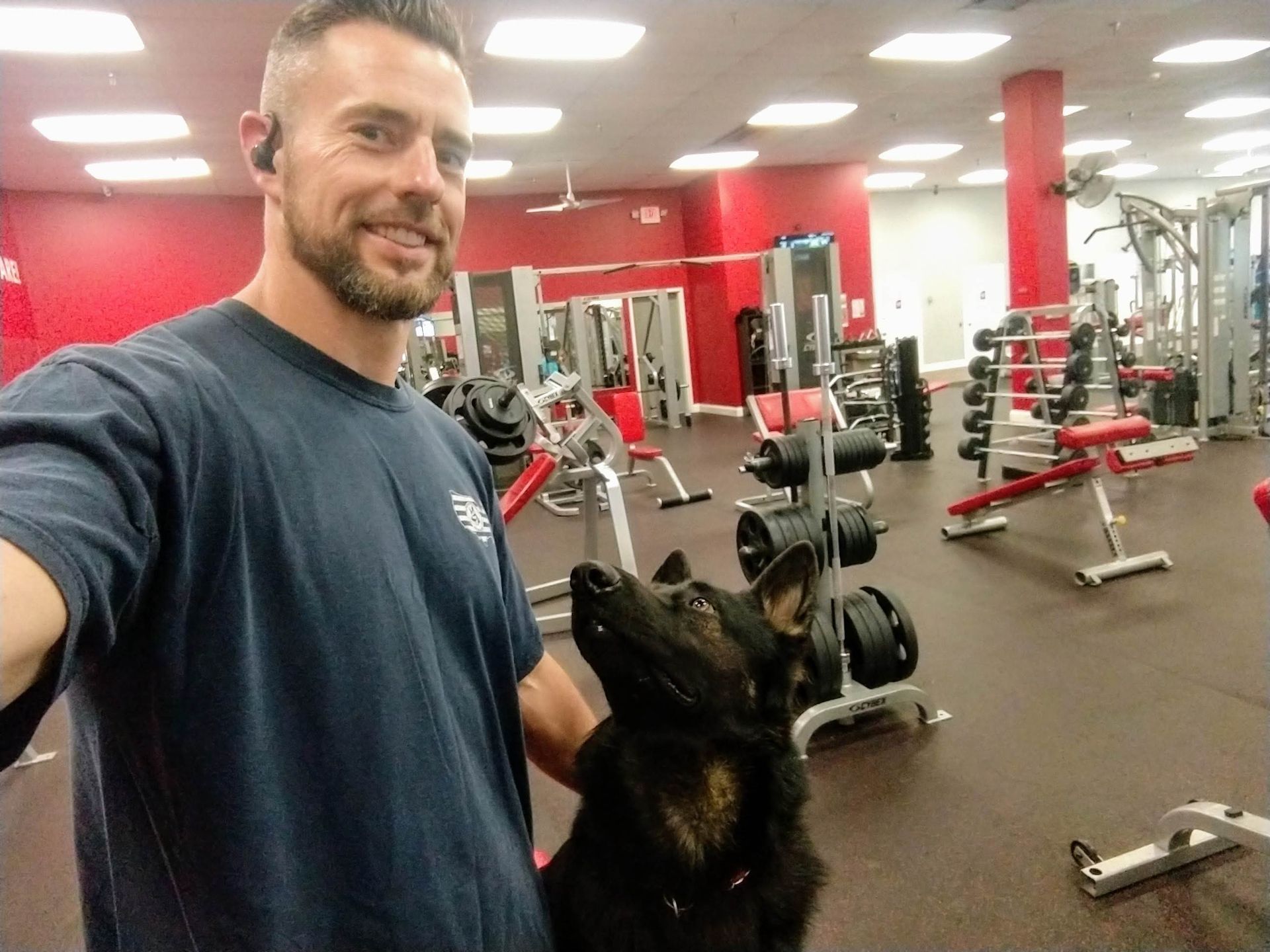 Man in gym takes selfie with a black dog. Red walls, weights, fitness equipment. Man smiles.