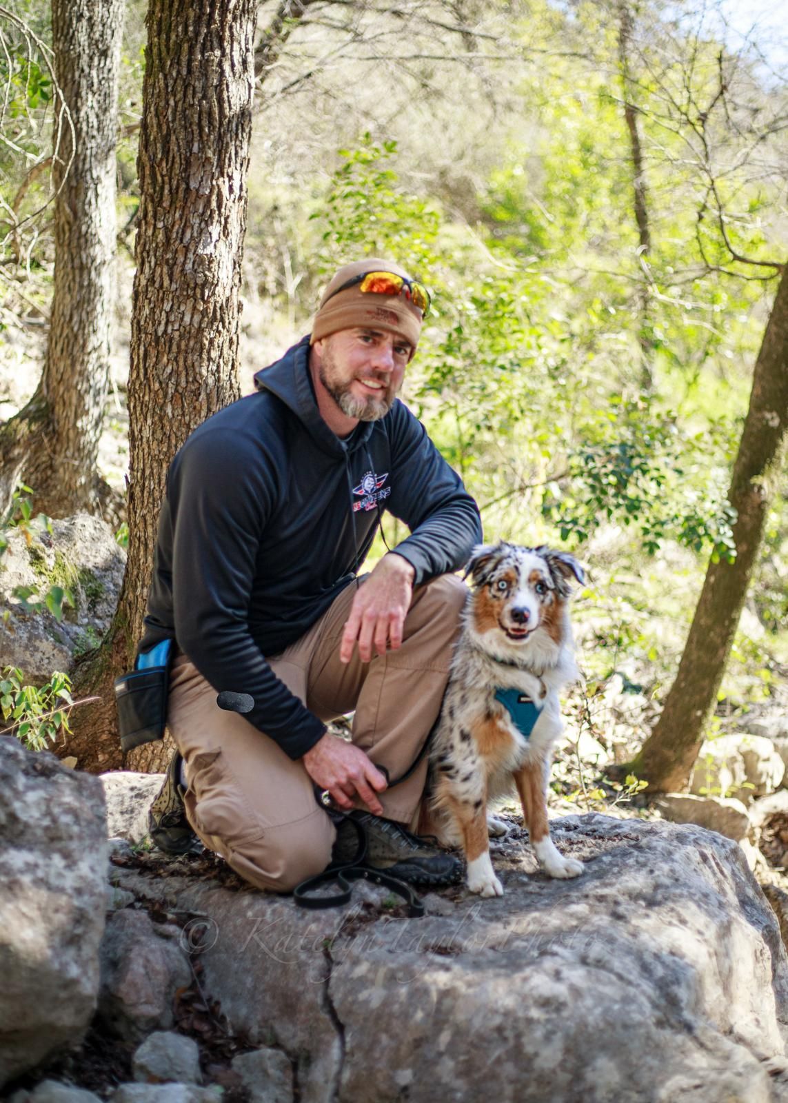 Man kneeling with a blue merle dog on a rock in a forest.
