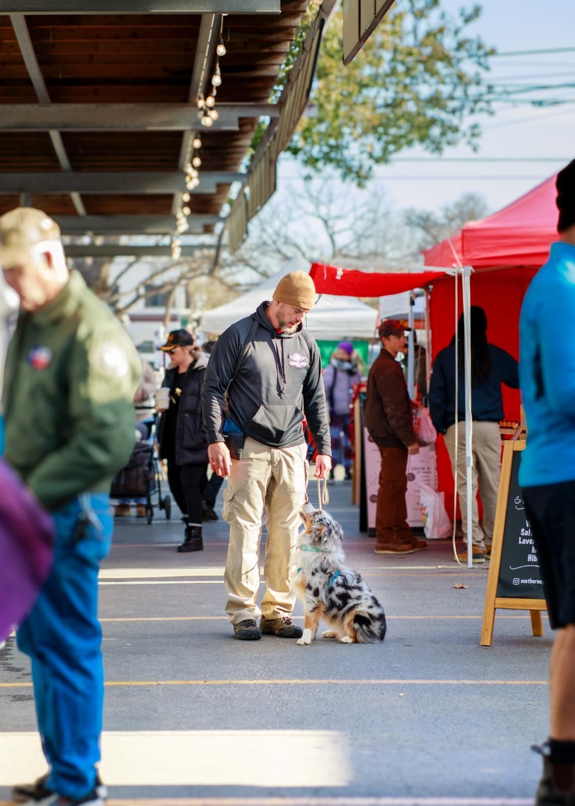 Man with dog at outdoor market. Dog is blue merle, man wears a black jacket and tan pants.